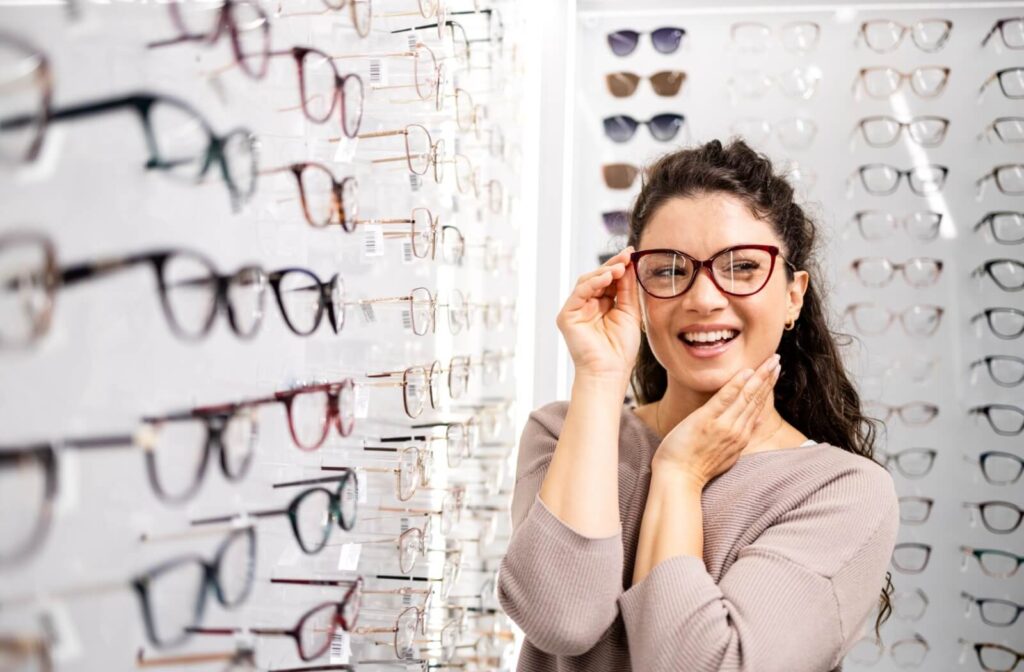 Person trying on eyeglasses in an optical store, smiling while standing in front of a wall display of frames.