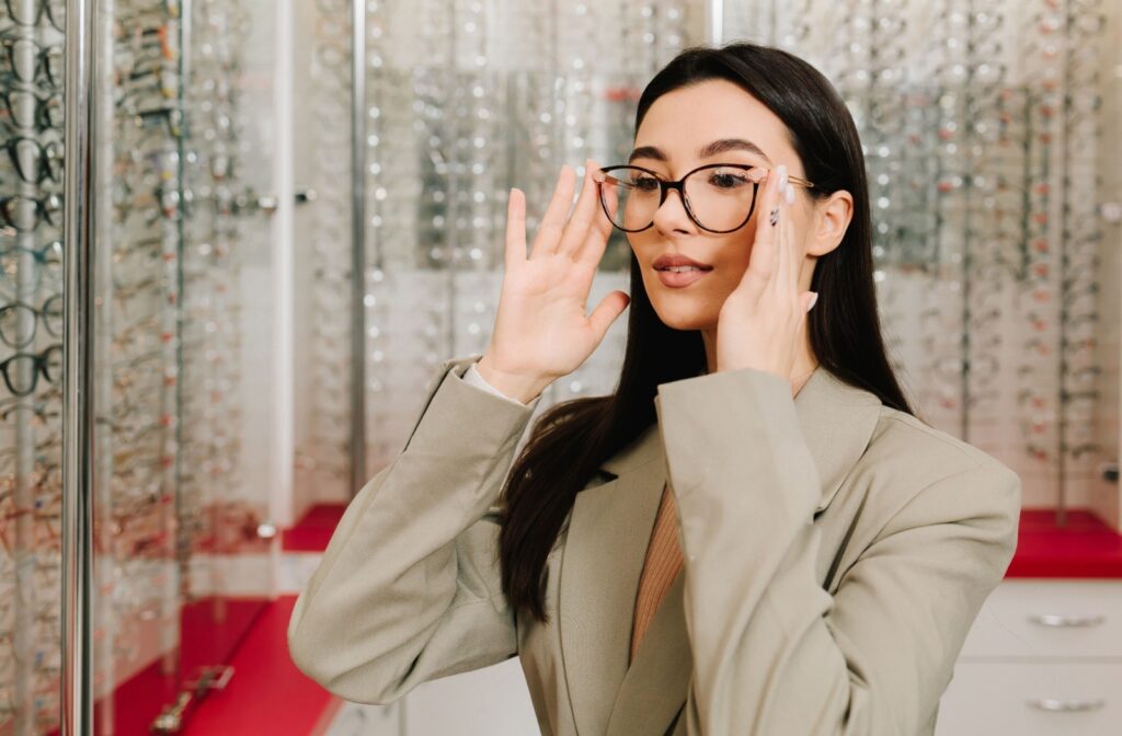 Person adjusting eyeglasses while standing inside an optical store, with rows of eyeglass frames displayed behind her.