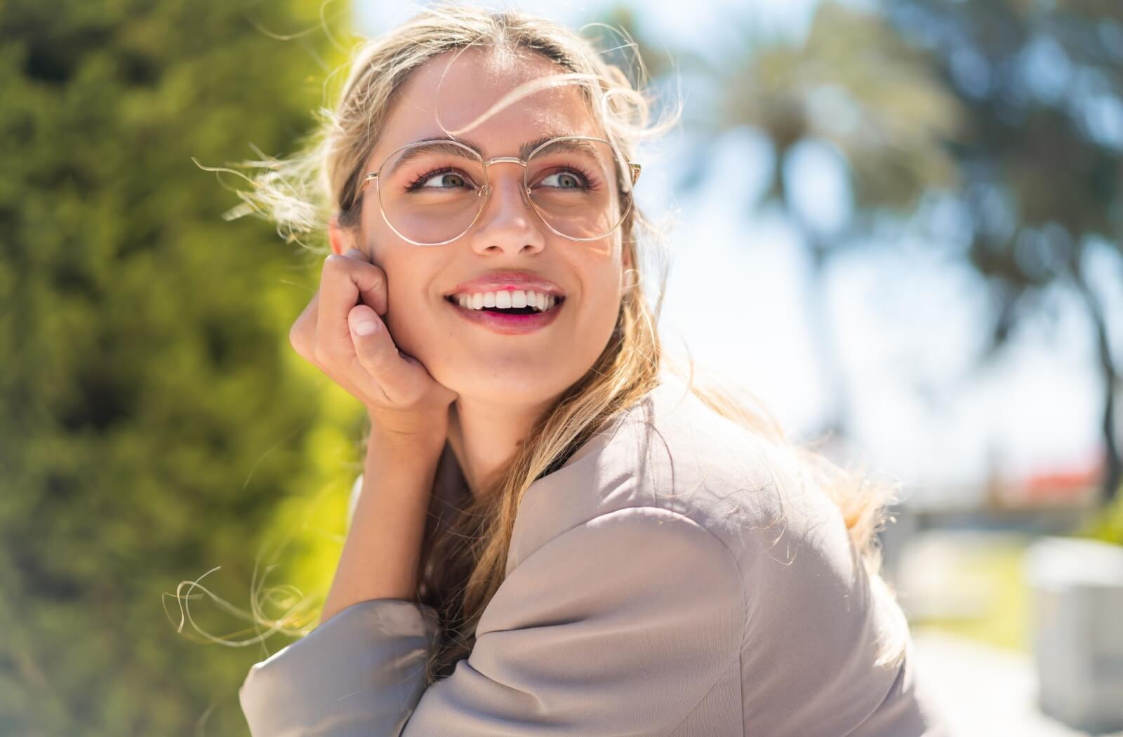 A smiling individual wearing oversized, round metal-rimmed eyeglasses while posing outdoors.